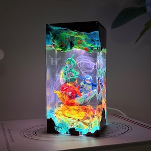 K&ouml;nnte beinhalten: Eine rechteckige Kunstharzlampe mit einem farbenfrohen Diorama aus drei kleinen Figuren, die in transparentes Material eingegossen sind. Der Sockel und die Oberseite sind dunkel, w&auml;hrend das Innere in hellem Gr&uuml;n, Blau und Orange leuchtet.