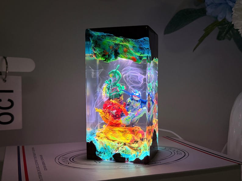 K&ouml;nnte beinhalten: Eine rechteckige Kunstharzlampe mit einem farbenfrohen Diorama aus drei kleinen Figuren, die in transparentes Material eingegossen sind. Der Sockel und die Oberseite sind dunkel, w&auml;hrend das Innere in hellem Gr&uuml;n, Blau und Orange leuchtet.