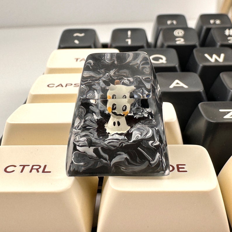 K&ouml;nnte beinhalten: Eine schwarz-wei&szlig;e marmorierte Tastenkappe mit einer durchsichtigen Oberseite. Die Tastenkappe hat eine kleine wei&szlig;e Figur im Inneren mit schwarzen Augen und einer gelben Nase.