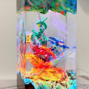K&ouml;nnte beinhalten: Eine rechteckige Lampe aus klarem Kunstharz mit drei Miniaturfiguren: ein gr&uuml;nes schlangenartiges Wesen, ein rotes gepanzertes Wesen und ein blaues walartiges Wesen. Die Basis zeigt wirbelnde orange, gelbe und blaue Farben wie Lava und Wasser.