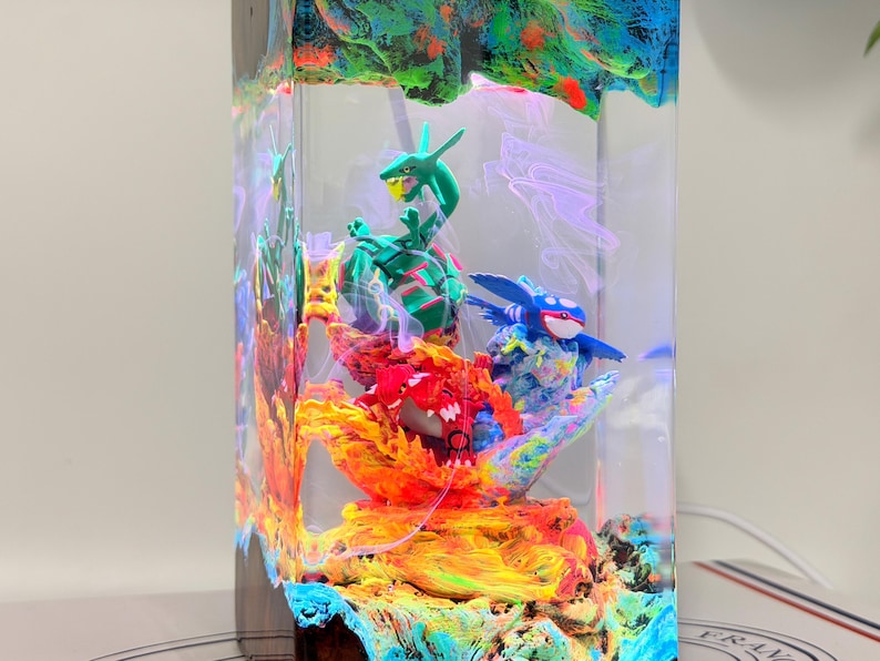 K&ouml;nnte beinhalten: Eine rechteckige Lampe aus klarem Kunstharz mit drei Miniaturfiguren: ein gr&uuml;nes schlangenartiges Wesen, ein rotes gepanzertes Wesen und ein blaues walartiges Wesen. Die Basis zeigt wirbelnde orange, gelbe und blaue Farben wie Lava und Wasser.