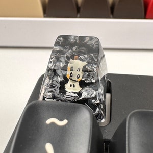 K&ouml;nnte beinhalten: Eine transparente, schwarz-wei&szlig;e marmorierte Tastaturkappe mit einem kleinen, wei&szlig;en Kuh-Design im Inneren. Die Kappe sitzt auf einer schwarzen Tastatur.