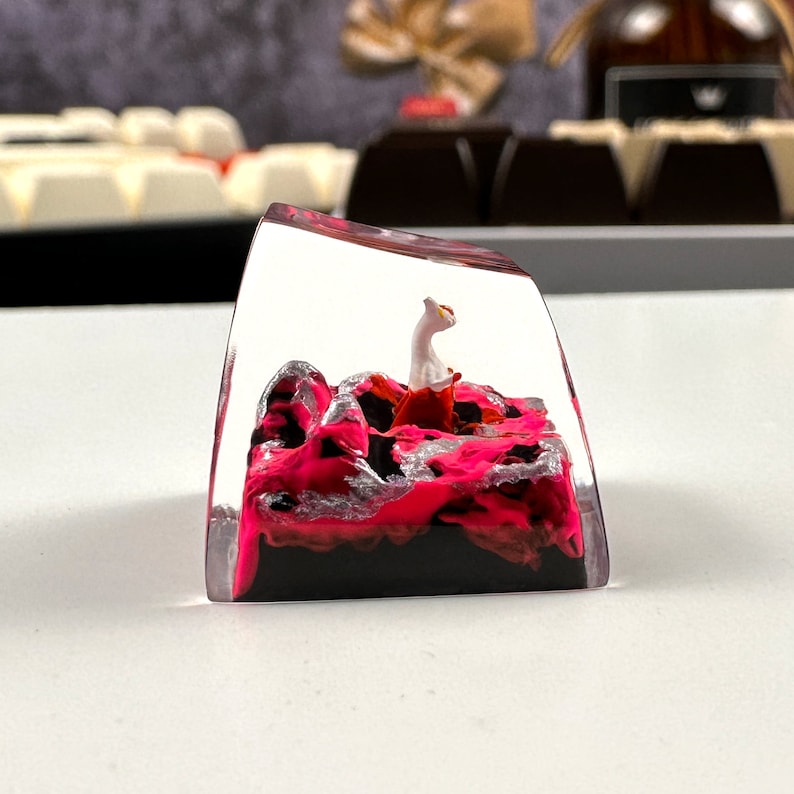 Pode incluir: Um keycap de resina transparente com um design de redemoinho rosa e preto. Uma pequena figura branca de um gato est&aacute; sentada no topo do redemoinho.