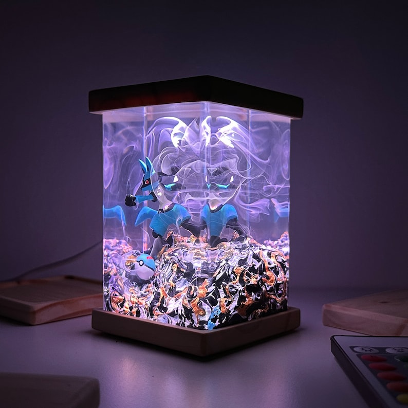 May include: A wooden lamp base with a clear glass enclosure containing two blue and white Pokemon figures, a Pokeball, and white smoke-like resin. The lamp is lit with a purple glow.