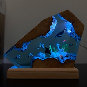 May include: A wooden sculpture with a clear resin insert depicting a blue ocean scene with a manta ray, fish, and sea turtles. The resin is illuminated from within, creating a glowing effect.