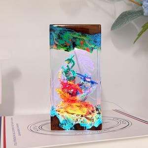 K&ouml;nnte beinhalten: Eine rechteckige Harzlampe mit einem Sockel und einer Oberseite aus dunklem Holz, die ein lebhaftes, mehrfarbiges Diorama mit drei kleinen Figuren umschlie&szlig;t. Die Szene im Inneren ist hell beleuchtet und zeigt gr&uuml;ne, blaue und rote Elemente.