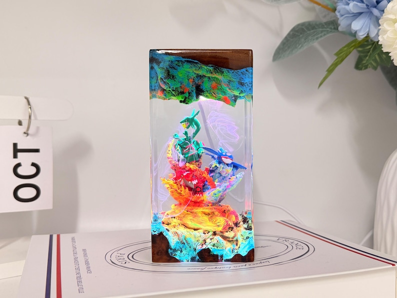 K&ouml;nnte beinhalten: Eine rechteckige Harzlampe mit einem Sockel und einer Oberseite aus dunklem Holz, die ein lebhaftes, mehrfarbiges Diorama mit drei kleinen Figuren umschlie&szlig;t. Die Szene im Inneren ist hell beleuchtet und zeigt gr&uuml;ne, blaue und rote Elemente.