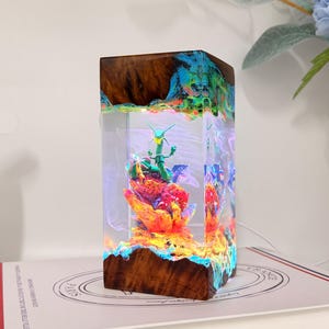 K&ouml;nnte beinhalten: Eine rechteckige Lampe aus dunklem Holz und klarem Harz mit einer gr&uuml;nen drachenartigen Figur auf einem leuchtend orangefarbenen und roten Felsensockel. Die innere Struktur ist mit bunten LED-Lichtern beleuchtet, was einen leuchtenden Effekt erzeugt.