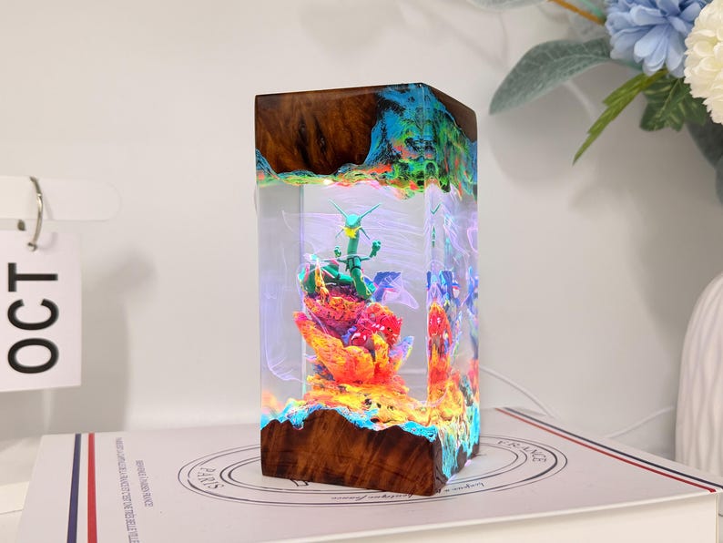 K&ouml;nnte beinhalten: Eine rechteckige Lampe aus dunklem Holz und klarem Harz mit einer gr&uuml;nen drachenartigen Figur auf einem leuchtend orangefarbenen und roten Felsensockel. Die innere Struktur ist mit bunten LED-Lichtern beleuchtet, was einen leuchtenden Effekt erzeugt.