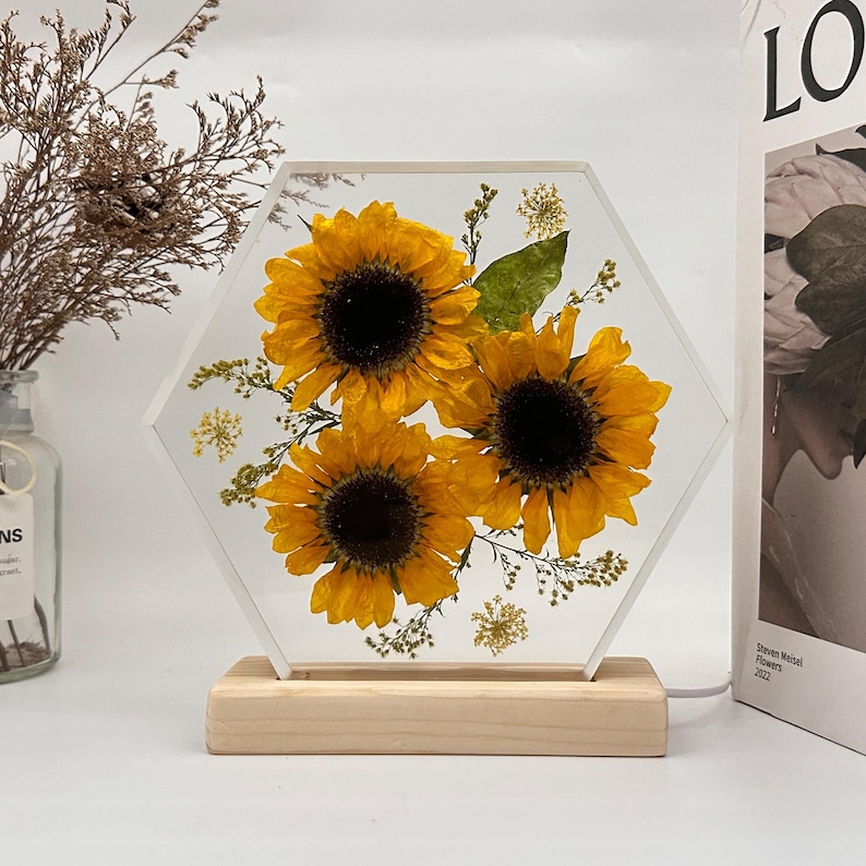 Pu&ograve; includere: Un blocco acrilico esagonale trasparente contenente tre girasoli gialli pressati e piccolo fogliame verde, montato su una base rettangolare in legno chiaro. L'oggetto presenta un cavo di alimentazione bianco che esce dal lato destro del supporto.