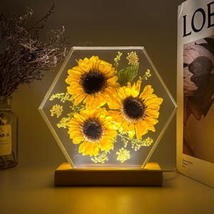 Pu&ograve; includere: Una lampada esagonale in resina trasparente contenente tre girasoli gialli pressati e piccoli fiori verdi di riempimento, illuminata dall'interno. Il pannello in resina &egrave; montato su una base rettangolare in legno chiaro, creando un display floreale luminoso.