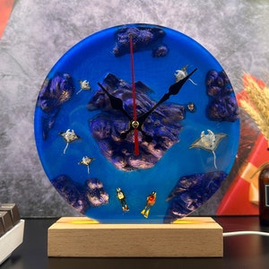 May include: A blue resin clock with a wooden base. The clock face features a purple and blue ocean scene with white manta rays and gold glitter. The clock hands are black and red.