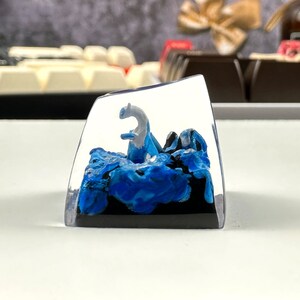 Pode incluir: Um keycap de resina transparente com uma figura branca e azul de uma criatura m&iacute;tica em p&eacute; sobre uma onda azul e preta. O keycap &eacute; um item decorativo para um teclado.