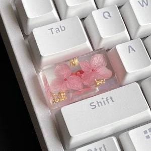 K&ouml;nnte beinhalten: Eine rosafarbene und goldene Harz-Tastenkappe mit gepressten Blumen. Die Tastenkappe befindet sich auf einer wei&szlig;en Tastatur mit den sichtbaren Tasten "Tab", "Q", "A", "Shift".
