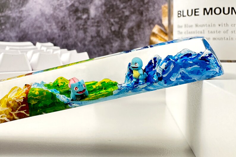 May include: A clear resin keycap with a painted landscape scene featuring blue water, green land, and two small blue and pink figurines.