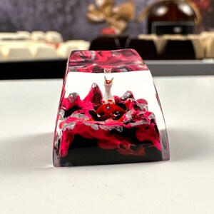 Pode incluir: Um keycap de resina transparente com um design de redemoinho rosa e preto. Uma pequena figura branca e vermelha est&aacute; dentro do keycap.