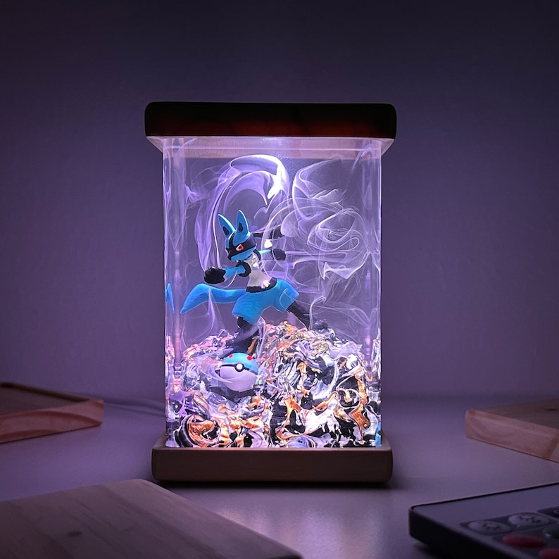 May include: A blue and black Pokemon character figurine is encased in a clear resin cube with a wooden base. The cube is illuminated from the inside, creating a glowing effect. The figurine is surrounded by gold and silver flakes.