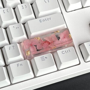 K&ouml;nnte beinhalten: Eine wei&szlig;e Tastatur mit einer rosafarbenen und goldenen Harz-Tastenkappe mit gepressten Blumen. Die Tastenkappe befindet sich auf der Taste "Strg".