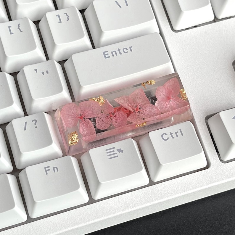 K&ouml;nnte beinhalten: Eine wei&szlig;e Tastatur mit einer rosafarbenen und goldenen Harz-Tastenkappe mit gepressten Blumen. Die Tastenkappe befindet sich auf der Taste "Strg".