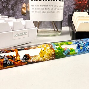 May include: A clear resin keyboard wrist rest with a multi-colored design featuring four different Pokemon characters. The wrist rest is divided into four sections, each with a different color and design. The first section is orange and black with a Charmeleon Pokemon character. The second section is yellow and white with a Pikachu Pokemon character. The third section is green and brown with a Bulbasaur Pokemon character. The fourth section is blue and white with a Squirtle Pokemon character.