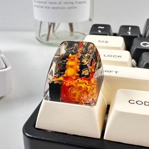 May include: A clear resin keycap with a small orange and yellow figurine of a Pokemon character inside. The resin is filled with swirls of orange, yellow, and black. The keycap is sitting on a white keycap base.