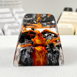 May include: A custom artisan keycap with a fiery orange creature set in clear resin. The keycap has black and orange accents, with a detailed design of a volcano and lava.