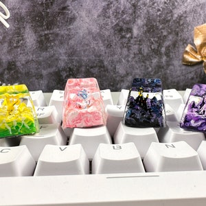 Könnte beinhalten: Vier individuelle Tastenkappen mit einzigartigen Designs. Eine zeigt einen gelben Charakter, eine andere ein rosa-weißes Muster, eine dritte eine dunkle Landschaft und die letzte eine lila Landschaft. Sie befinden sich auf einer weißen Tastatur.