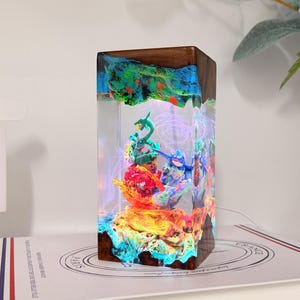 K&ouml;nnte beinhalten: Eine rechteckige Harz- und Holzlampe mit einem klaren Mittelteil und interner LED-Beleuchtung. Im Inneren des Harzes befinden sich bunte, geformte Figuren in Gr&uuml;n, Blau und Rot. Die oberen und unteren Kanten bestehen aus dunkelbraunem Holz.