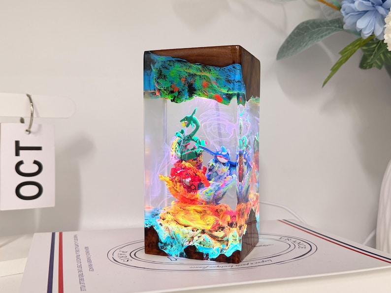 K&ouml;nnte beinhalten: Eine rechteckige Harz- und Holzlampe mit einem klaren Mittelteil und interner LED-Beleuchtung. Im Inneren des Harzes befinden sich bunte, geformte Figuren in Gr&uuml;n, Blau und Rot. Die oberen und unteren Kanten bestehen aus dunkelbraunem Holz.