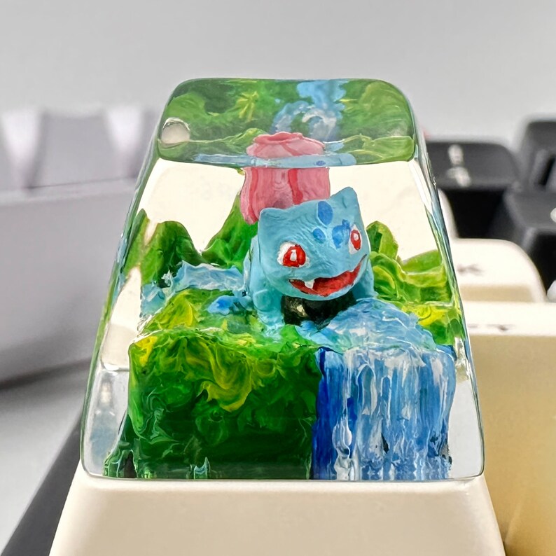 May include: A clear resin keycap with a green, blue, and white landscape scene. A small blue and white figurine of a Pokemon character is standing on a green hill with a waterfall behind it. A pink flower is partially visible behind the figurine.