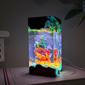 K&ouml;nnte beinhalten: Ein rechteckiges Nachtlicht aus Kunstharz mit einer gr&uuml;nen, drachen&auml;hnlichen Figur auf einem leuchtend orangefarbenen und roten, korallenartigen Sockel. Der klare Kunstharzk&ouml;rper enth&auml;lt wirbelnde wei&szlig;e Muster und wird von innen durch LEDs beleuchtet.