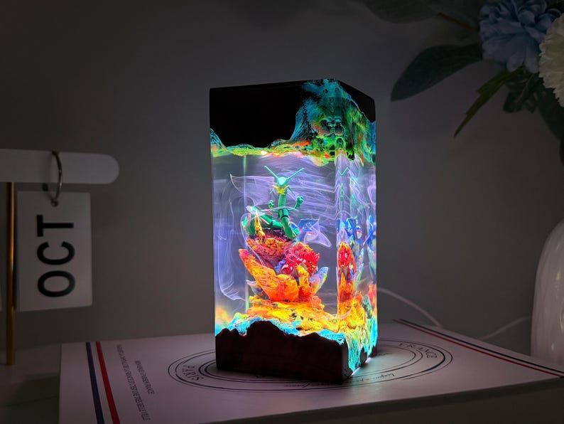 K&ouml;nnte beinhalten: Ein rechteckiges Nachtlicht aus Kunstharz mit einer gr&uuml;nen, drachen&auml;hnlichen Figur auf einem leuchtend orangefarbenen und roten, korallenartigen Sockel. Der klare Kunstharzk&ouml;rper enth&auml;lt wirbelnde wei&szlig;e Muster und wird von innen durch LEDs beleuchtet.