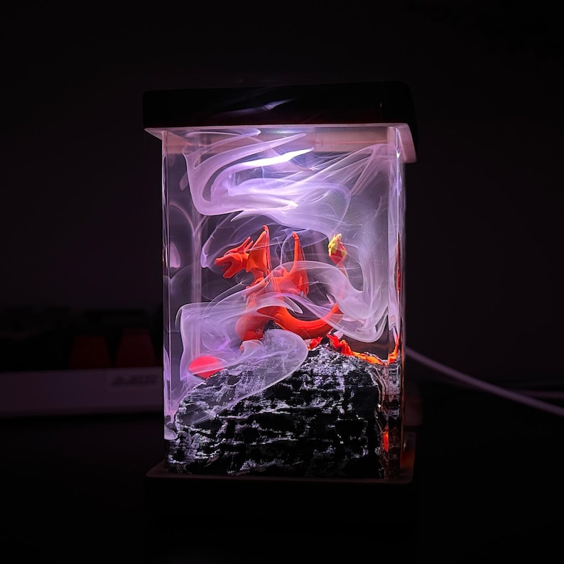 K&ouml;nnte beinhalten: Eine rote Drachenfigur, die in klarem Harz mit wei&szlig;en Wirbeln eingeschlossen ist und von einem violetten Licht beleuchtet wird. Der Drache sitzt auf einem schwarzen Felssockel.