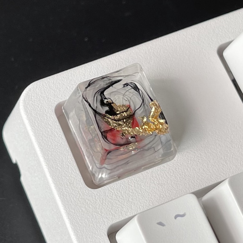 K&ouml;nnte beinhalten: Eine transparente Harztastenkappe mit schwarzen und roten abstrakten Wirbeln und goldenen Flocken. Die Tastenkappe ist f&uuml;r eine mechanische Tastatur konzipiert.