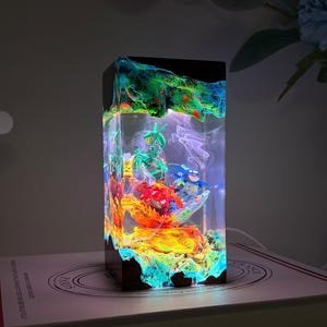K&ouml;nnte beinhalten: Eine rechteckige Harzlampe mit einem Sockel und einer Oberseite aus dunklem Holz, die eine Miniaturszene mit bunten Figuren umschlie&szlig;t, darunter ein gr&uuml;nes, ein blaues und ein rotes Wesen, beleuchtet von einem hellen wei&szlig;en Licht im Inneren.