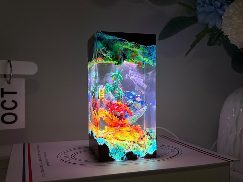 K&ouml;nnte beinhalten: Eine rechteckige Harzlampe mit einem Sockel und einer Oberseite aus dunklem Holz, die eine Miniaturszene mit bunten Figuren umschlie&szlig;t, darunter ein gr&uuml;nes, ein blaues und ein rotes Wesen, beleuchtet von einem hellen wei&szlig;en Licht im Inneren.