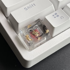 K&ouml;nnte beinhalten: Eine transparente Harztastenkappe mit schwarzen und goldenen Wirbeln und einem roten Akzent. Die Tastenkappe liegt auf einer wei&szlig;en Tastatur.
