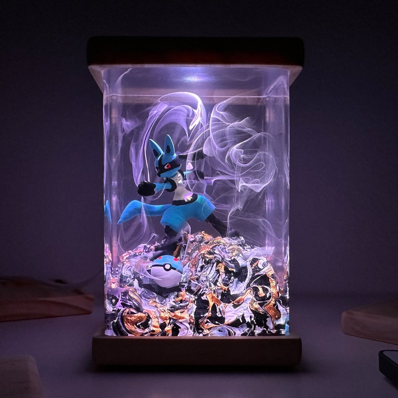 May include: A blue and white Pokemon figurine of Lucario is encased in a clear resin cube with a swirling white design. The cube is lit from the bottom with a white light. The figurine is standing on a bed of gold and black glitter.