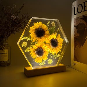 Pu&ograve; includere: Un blocco di resina trasparente esagonale contenente tre girasoli gialli conservati e piccoli fiori verdi, illuminato dal basso da una base in legno rettangolare. L'oggetto presenta petali gialli vivaci e centri marrone scuro.