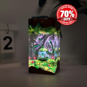 May include: A rectangular resin lamp featuring a light-green Bulbasaur figure surrounded by green moss, small red mushrooms, and swirling purple smoke effects, all encased in a clear block with dark wood-toned top and bottom sections.