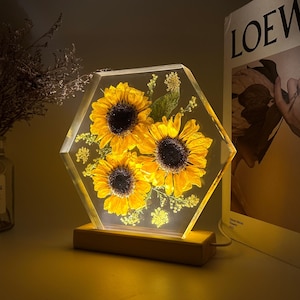 Pu&ograve; includere: Un blocco esagonale in resina trasparente contenente tre girasoli gialli pressati e del piccolo fogliame verde, illuminato dall'interno. L'oggetto in resina &egrave; montato su una base rettangolare in legno chiaro con un cavo di alimentazione laterale.