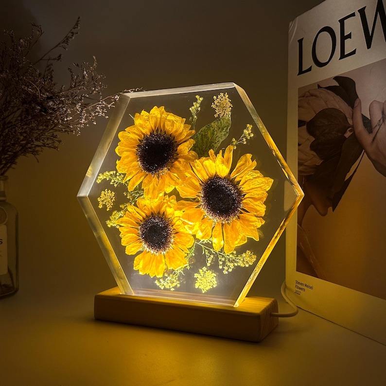 Pu&ograve; includere: Un blocco esagonale in resina trasparente contenente tre girasoli gialli pressati e del piccolo fogliame verde, illuminato dall'interno. L'oggetto in resina &egrave; montato su una base rettangolare in legno chiaro con un cavo di alimentazione laterale.