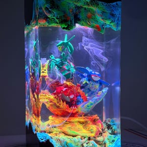 K&ouml;nnte beinhalten: Eine rechteckige Harzlampe mit einem transparenten Block, der drei Miniaturfiguren umschlie&szlig;t: ein gr&uuml;nes schlangenartiges Wesen, ein blaues gefl&uuml;geltes Wesen und ein rotes gepanzertes Wesen. Basis und Oberseite sind in leuchtenden Farben texturiert.