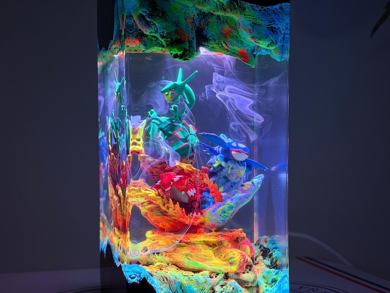 K&ouml;nnte beinhalten: Eine rechteckige Harzlampe mit einem transparenten Block, der drei Miniaturfiguren umschlie&szlig;t: ein gr&uuml;nes schlangenartiges Wesen, ein blaues gefl&uuml;geltes Wesen und ein rotes gepanzertes Wesen. Basis und Oberseite sind in leuchtenden Farben texturiert.