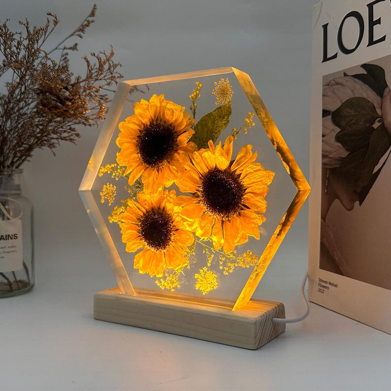 Pu&ograve; includere: Una lampada esagonale in resina trasparente contenente tre girasoli gialli conservati e del piccolo fogliame verde. L'elemento in resina &egrave; illuminato dall'interno e montato su una base rettangolare in legno chiaro dotata di cavo di alimentazione.