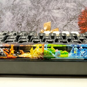 May include: A keyboard with a colorful resin keycap featuring a Pokemon theme. The keycap is divided into four sections, each with a different color and Pokemon character. The first section is orange and features a Charmander, the second is yellow and features a Pikachu, the third is green and features a Bulbasaur, and the fourth is blue and features a Squirtle.