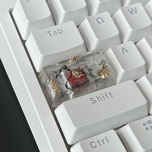 K&ouml;nnte beinhalten: Eine wei&szlig;e Tastatur mit einer benutzerdefinierten Tastenkappe. Die Tastenkappe besteht aus klarem Harz mit schwarzen und goldenen Akzenten. Die Tastenkappe hat einen roten Akzent in der Mitte.