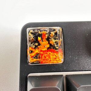 May include: A clear resin keycap with a gold and orange Pokemon character inside. The character is surrounded by a swirling orange and yellow design.