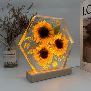 Pu&ograve; includere: Un blocco di resina trasparente esagonale contenente tre girasoli gialli pressati e piccolo fogliame verde, illuminato dall'interno. Il pezzo in resina &egrave; montato su una base rettangolare in legno chiaro con cavo di alimentazione.