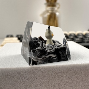 K&ouml;nnte beinhalten: Ein transparenter Harz-Tastenkappen mit einem schwarzen und wei&szlig;en Wirbelmuster. Eine kleine wei&szlig;e Figur eines Katzes befindet sich im Inneren der Tastenkappe.